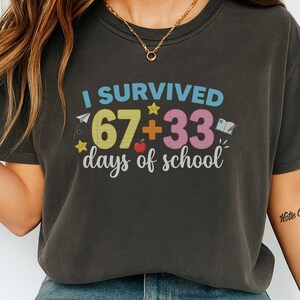 Puede incluir: Camiseta gris oscuro con el texto "I Survived 67+33 days of school" en letras coloridas. El diseño incluye un avión de papel, estrellas, una manzana y un libro. La camiseta es de estilo casual y cómodo.