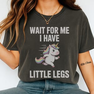 Puede incluir: Camiseta gris oscuro con el texto "WAIT FOR ME I HAVE LITTLE LEGS" arriba y abajo de un unicornio de dibujos animados con melena y cola arcoíris. El unicornio está corriendo. La camiseta es de estilo casual.