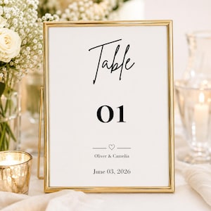 May include: A framed table number sign with a gold border. The sign has the word "Table" in a script font, the number "01", names "Oliver & Camelia", and the date "June 03, 2026".