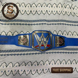 May include: A blue and gold WWE Universal Championship belt. The belt features a large, central plate with the WWE logo and the words "UNIVERSAL CHAMPION". The strap is blue with gold accents and the belt is on a white patterned surface.