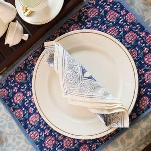 Puede incluir: Un plato blanco con borde dorado y una servilleta blanca doblada con un diseño floral azul se encuentra sobre un mantel individual azul. El mantel individual tiene un estampado floral con flores rosas. Una bandeja de madera con tazas de té blancas está en el fondo.
