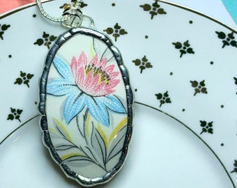 May include: Oval pendant necklace with a floral design. The pendant features a pink and blue flower on a white background, framed by a silver-colored metal. The necklace is on a silver chain, resting on a white plate with a floral pattern.