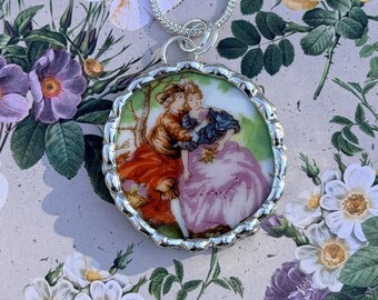 May include: Silver-toned pendant necklace featuring a round, hand-painted porcelain cameo. The cameo depicts two figures in period clothing, set against a green and brown backdrop. The pendant is suspended from a delicate silver chain.