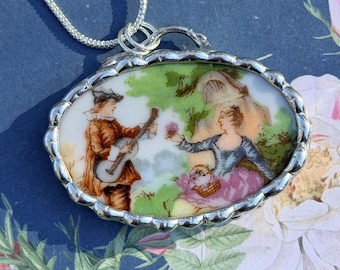 May include: Oval pendant necklace with a silver-colored frame. The pendant features a painted scene of a man playing a guitar and a woman with a basket. The scene is set in a garden with green foliage and a white background. The necklace chain is silver.