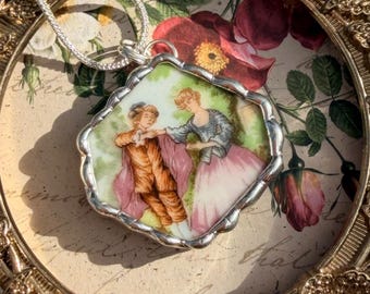 May include: Silver-toned pendant necklace featuring a rectangular, scalloped-edge pendant with a painted scene of two figures in period clothing. The pendant is set against a floral background with a silver chain.