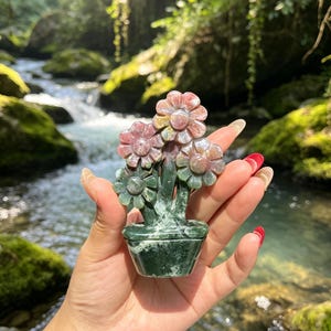 May include: A carved stone sculpture of three flowers in a pot. The flowers have pink and green petals, while the stems and the pot are a deep, solid green. The sculpture is crafted from a single piece of polished mineral with natural color variations.