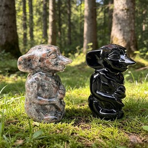 May include: Two stone sculptures of whimsical figures with large noses and folded arms. One is a speckled gray and pink, the other is glossy black. They are set on green grass with a forest background.