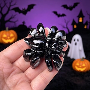 May include: A glossy black spider figurine with eight legs. The spider is held in a hand against a Halloween-themed backdrop with pumpkins, a ghost, and a haunted house. The background is purple.