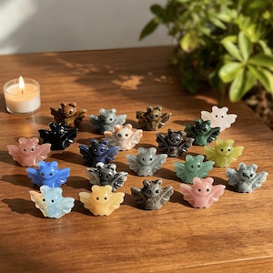 Puede incluir: Una colecci&oacute;n de peque&ntilde;as figuras de murci&eacute;lagos de piedra tallada en varios colores, incluyendo rosa, azul, verde y marr&oacute;n, est&aacute;n dispuestas sobre una superficie de madera. Una vela encendida est&aacute; en el fondo. Los murci&eacute;lagos tienen alas extendidas y detalles.