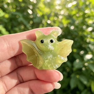 Puede incluir: Una peque&ntilde;a figura de murci&eacute;lago verde claro con ojos negros y alas extendidas. El murci&eacute;lago est&aacute; hecho de un material transl&uacute;cido, posiblemente una piedra preciosa, y se sostiene en una mano sobre un fondo verde borroso.