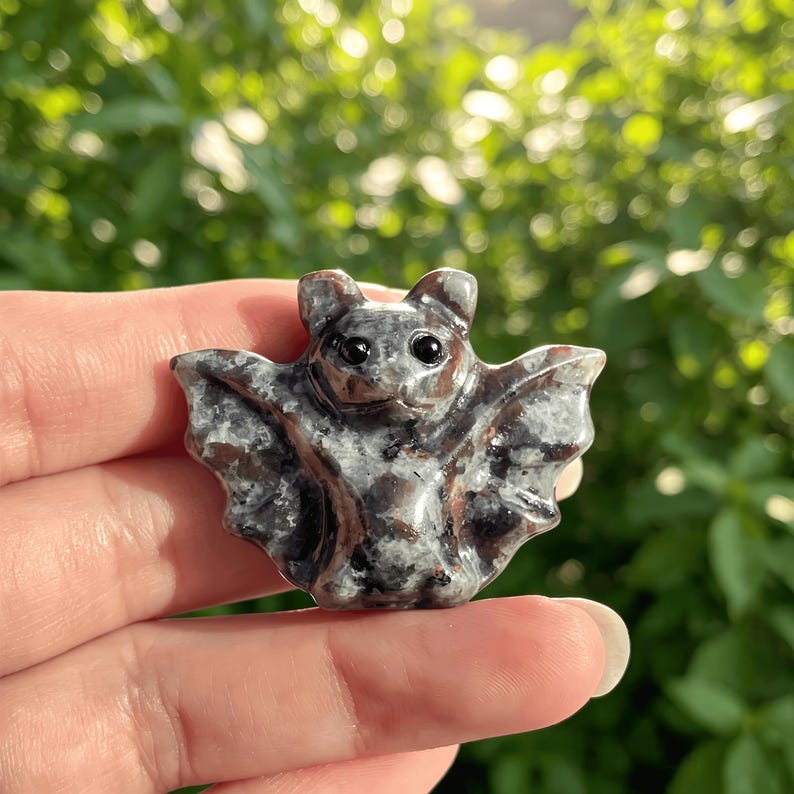 Puede incluir: Peque&ntilde;a figura de murci&eacute;lago de piedra tallada. El murci&eacute;lago es principalmente gris con vetas marrones y negras, y tiene ojos peque&ntilde;os y oscuros. Las alas est&aacute;n extendidas y la forma general es compacta y detallada.