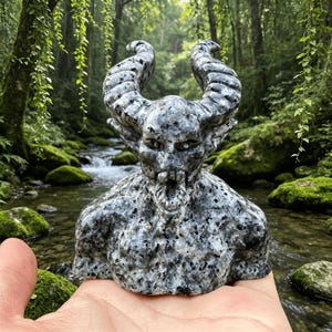 May include: A carved stone bust of a horned creature with a speckled black, grey, and white pattern. The figure features a wide, open mouth, defined facial features, and two large, curved horns extending upwards from the top of the head.
