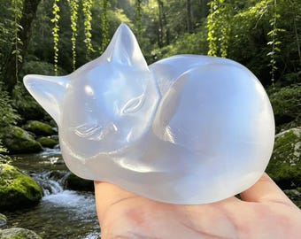 May include: A translucent, milky white, smooth stone carving of a curled-up sleeping cat. The sculpture features soft, rounded contours and subtle etched lines defining the closed eyes, ears, and body shape, reflecting light with a soft glow.