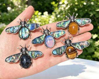 Puede incluir: Cinco broches con forma de abeja con cuerpos de metal plateado y alas de concha de abulón iridiscente. Cada broche tiene una piedra central distinta: una azul oscuro, una blanca translúcida, una ámbar, una ojo de tigre y una gris oscuro.