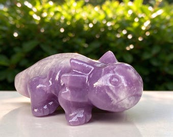 Puede incluir: Una figura de piedra tallada con forma de pequeño lagarto o dragón, que presenta una superficie lisa y pulida con una coloración jaspeada en violeta claro y blanco, rasgos redondeados y una cresta texturizada en la cabeza y el lomo.