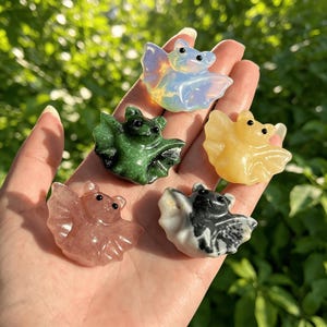 Puede incluir: Cinco figuritas de murci&eacute;lagos de piedras preciosas talladas en varios colores, incluyendo verde, rosa, amarillo, opalino y blanco y negro. Los murci&eacute;lagos tienen alas detalladas y ojos peque&ntilde;os y oscuros. El fondo es follaje verde borroso.