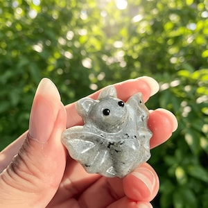 Puede incluir: Figurita de murci&eacute;lago de piedra gris con ojos negros y alas detalladas. El murci&eacute;lago se sostiene en una mano sobre un fondo verde borroso. La piedra tiene un patr&oacute;n moteado y est&aacute; tallada con detalles.
