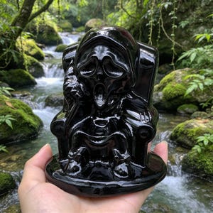 Könnte beinhalten: Eine polierte schwarze Obsidian-Schnitzerei, die eine Kapuzenfigur mit einer stilisierten schreienden Maske darstellt, die auf einem Stuhl sitzt. Die Figur hat ausgehöhlte Augenhöhlen und einen offenen Mund mit glatten, reflektierenden Oberflächen.
