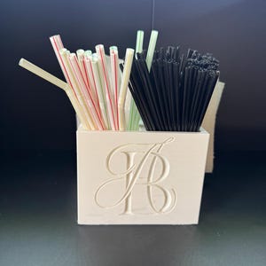 May include: A white square container with a large embossed letter B on the front, filled with a collection of drinking straws. The straws are divided into two sections, with one side containing white straws with red and green stripes and the other side containing solid black straws.