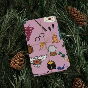 Può includere: Un regalo avvolto con uno sfondo rosa e un motivo ripetuto di simboli magici, tra cui occhiali, una bacchetta e un gufo che porta una lettera. Il regalo è legato con spago e poggia su un letto di aghi di pino verdi e pigne.