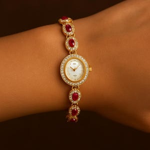 Puede incluir: Reloj de pulsera dorado con esfera blanca y bisel incrustado con pequeños cristales transparentes. La correa presenta piedras preciosas rojas ovaladas engastadas en oro. El reloj se muestra en la muñeca.