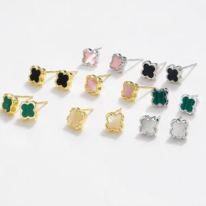 May include: A collection of clover-shaped stud earrings in various colors, including black, green, pink, white, and gold. The earrings are displayed on a white surface, showcasing their different designs and metallic finishes.