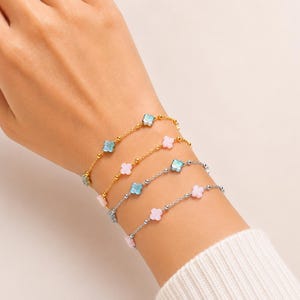 May include: Four delicate bracelets with clover-shaped charms in gold and silver tones. The charms are light blue and pink. The bracelets are worn on a wrist, showcasing the jewelry's design and color palette.