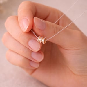 Puede incluir: Un delicado collar de oro con un cordón transparente, casi invisible. El colgante presenta tres anillos apilados, cada uno adornado con pequeños cristales brillantes. El collar se sostiene en una mano con uñas cuidadas, mostrando la sutil elegancia de la joya.