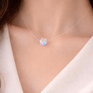 Puede incluir: Un delicado collar con un cordón transparente y un colgante de cubo iridiscente. El cubo refleja múltiples colores. El collar es un accesorio minimalista. El colgante es un pequeño cubo, creando un efecto sutil y llamativo.
