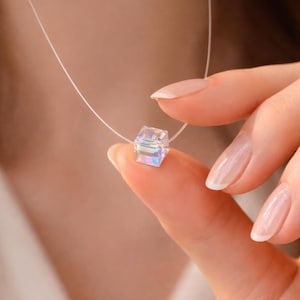 Puede incluir: Un delicado collar con un colgante cúbico transparente con reflejos iridiscentes. El colgante está suspendido de un cordón fino y casi invisible. El collar está sujeto por los dedos de una persona.