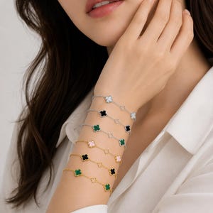 Peut inclure: Plusieurs bracelets avec des breloques en forme de trèfle de différentes couleurs, dont blanc, noir, vert et rose. Les bracelets sont en argent et en or, et sont portés au poignet. La personne porte une chemise blanche.