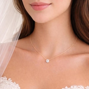 以下が含まれることがあります： 丸い、キラキラ光るペンダントが付いた繊細なゴールドネックレス。ペンダントは、小さな透明な石のクラスターです。ネックレスは人の首に着用され、背景には白いベールとレースが見えます。