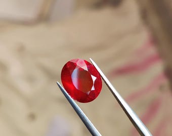 Pode incluir: Uma vibrante gema de rubi de corte oval, segurada por pinças prateadas. A gema exibe um tom vermelho profundo com facetas internas que refletem a luz. O fundo está desfocado, destacando a gema.
