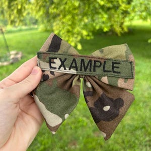 May include: A fabric hair bow featuring a camouflage pattern in shades of green, brown, and tan. A rectangular strip of olive green fabric is sewn across the center of the bow, with the word EXAMPLE embroidered in black capital letters.