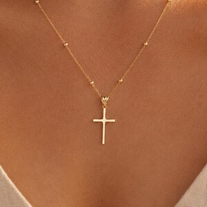 May include: A delicate gold necklace featuring a thin chain with evenly spaced small gold beads and a simple, smooth gold cross pendant hanging from the center. The necklace is worn around the neck, showcasing the polished metal finish and design.