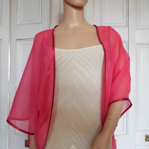 May include: A pink sheer shawl with a red satin trim. The shawl is draped over a white dress form.