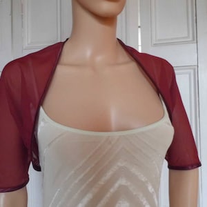 Op de afbeelding: Een bordeauxrode chiffon shrug met korte mouwen en een satijnen rand. De shrug is doorschijnend en lichtgewicht.