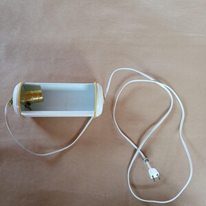 May include: A vintage, white, cylindrical lamp with yellow trim and a gold-colored bulb socket. The lamp is connected to a white electrical cord with a two-prong plug. The lamp is on a brown surface.