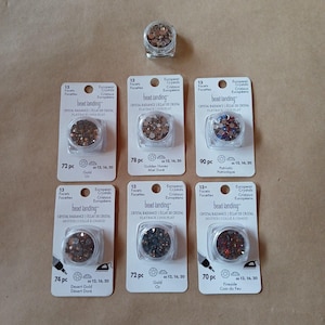 May include: An assortment of bead landing crystal rhinestones in various colors, including gold, brown, and red. The rhinestones are packaged in small, clear containers and attached to white cards. The cards have the text "bead landing" and "European Crystals".