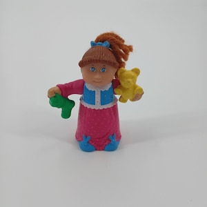 May include: A vintage doll with brown hair in a ponytail, blue eyes, and a blue bow. The doll wears a pink dress with blue accents and holds a green toy and a yellow teddy bear. A collectible toy.