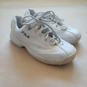 Puede incluir: Par de zapatillas deportivas Fila blancas con cordones. Las zapatillas tienen el logotipo de Fila en rojo y azul marino. Las suelas son blanquecinas y las zapatillas parecen ser de cuero.