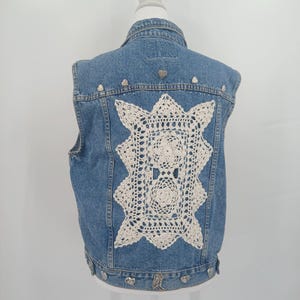 May include: A blue denim vest featuring a large, rectangular cream-colored crochet doily appliqué on the back. The vest is embellished with small, metallic heart-shaped studs along the upper back yoke and the bottom hemline.
