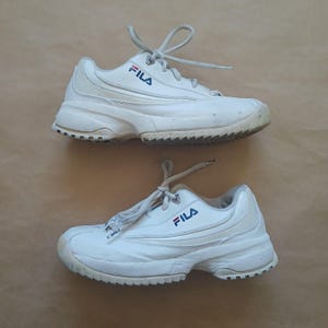 Puede incluir: Par de zapatillas deportivas Fila blancas con logo azul y rojo. Las zapatillas tienen cordones blancos y una suela gruesa y texturizada. Sobre una superficie marr&oacute;n claro.