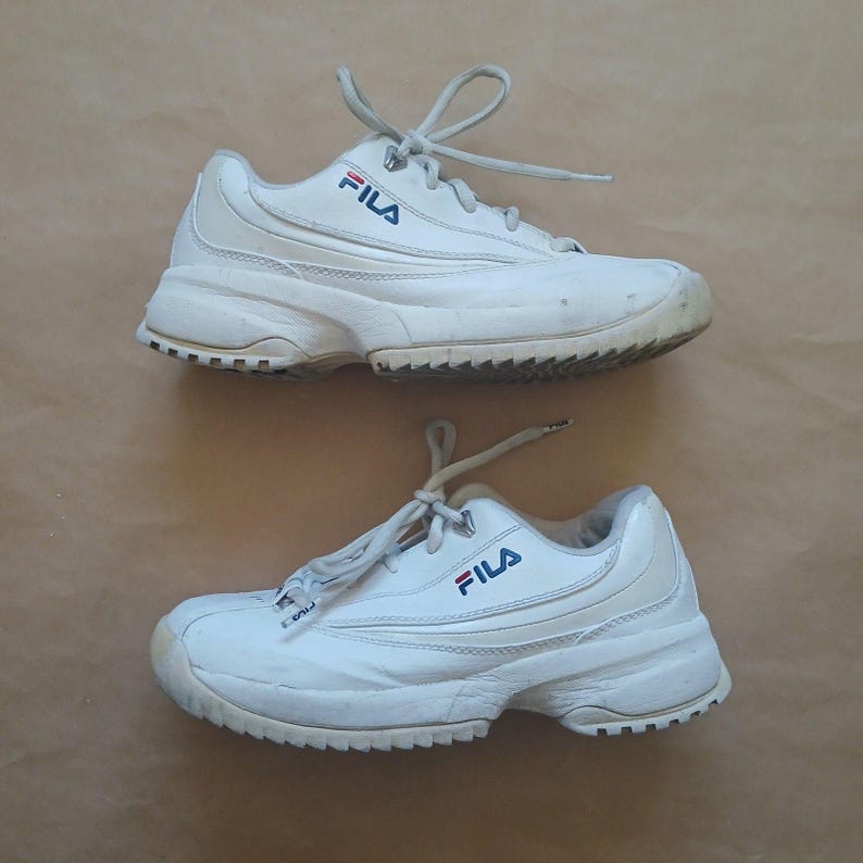 Puede incluir: Par de zapatillas deportivas Fila blancas con logo azul y rojo. Las zapatillas tienen cordones blancos y una suela gruesa y texturizada. Sobre una superficie marr&oacute;n claro.