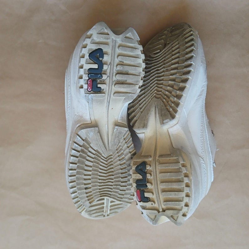Puede incluir: Zapatillas FILA blanquecinas con suela gruesa. Las suelas est&aacute;n sucias, mostrando desgaste. El logotipo de FILA es visible en negro y rojo en la suela. Las zapatillas est&aacute;n sobre una superficie marr&oacute;n.