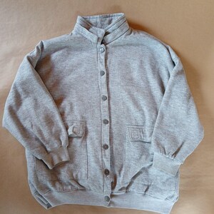 May include: A gray button-up sweatshirt with a high collar and two front pockets. The sweatshirt has long sleeves with ribbed cuffs and a button closure down the front. The fabric appears to be a soft, comfortable material.