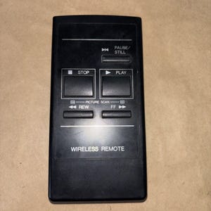 May include: A black wireless remote control with several buttons, including play, stop, rewind, and fast forward. The remote also has a pause/still button and the words "Wireless Remote" printed on the bottom.