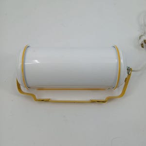 May include: A cylindrical white light fixture with a glossy finish, featuring bright yellow trim around the edges. The light is mounted on a yellow metal bracket and includes a white electrical cord with a standard two-prong plug attached.