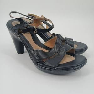 May include: Pair of black leather sandals with a high heel and ankle strap. The sandals have a platform sole and a decorative strap across the toes. The brand name "Born" is visible on the side of the shoe. The heels are approximately 3.5 inches high.