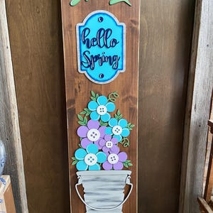 May include: Wooden sign with the words "hello spring" in blue lettering on a blue background. The sign also features a bucket with flowers in shades of blue, purple, and white.